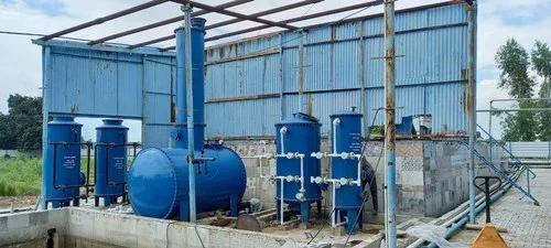 residential-sewage-treatment-plant Industrial water treatment setup with multiple blue cylindrical filtration tanks, connected piping, and a horizontal pressure vessel inside a partially open metal shed.