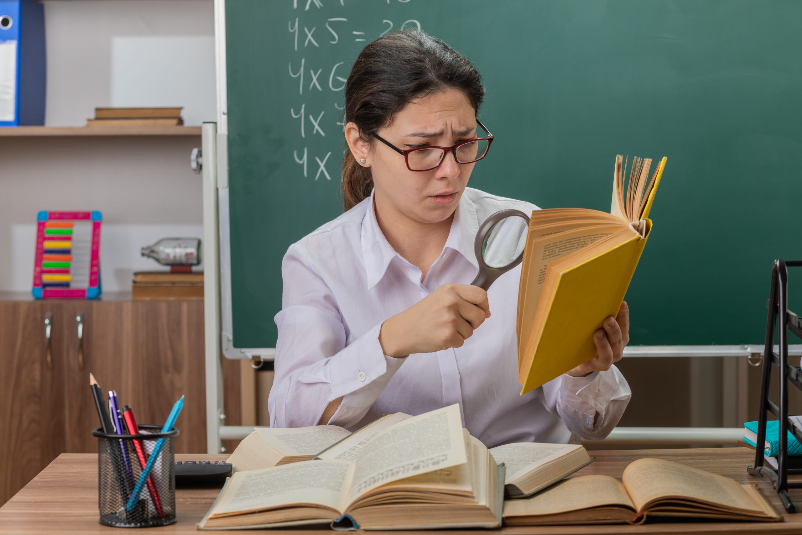 young woman teacher wearing glasses looking at book through magnifying glass being confused and displeased sitting at school desk in front of blackboard in classroom young woman teacher wearing glasses looking at book through magnifying glass being confused and displeased sitting at school desk in front of blackboard in classroom
